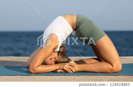 On windy day young slender woman does yoga near sea doing pigeon pose 118419865