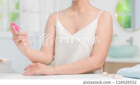 A model in a white camisole gently holds a spray bottle, ready to apply it to her skin. The soft lighting and calm atmosphere suggest a skincare or beauty routine in a serene bathroom setting. A model in a white camisole gently holds a spray bottle, ready to apply it to her skin. The soft lighting and calm atmosphere suggest a skincare or beauty routine in a serene bathroom setting. 118420552