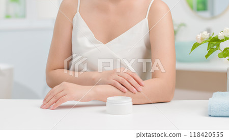 Cosmetic advertisement photo featuring woman sits at white table with a small jar of moisturizer in front of her to apply it in the bathroom background with soft natural lighting and minimalist decor 118420555