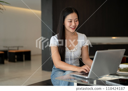 An attractive, smiling Asian woman working remotely from a coffee shop, working on her laptop. 118424187