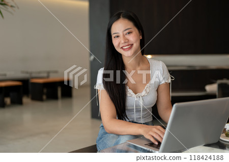 An attractive Asian woman working remotely from a cafe, working on her laptop, smiling at the camera An attractive Asian woman working remotely from a cafe, working on her laptop, smiling at the camera 118424188