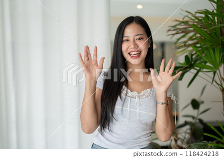 A cheerful Asian woman is standing by a window in a room, smiling and waving her hand at the camera. A cheerful Asian woman is standing by a window in a room, smiling and waving her hand at the camera. 118424218