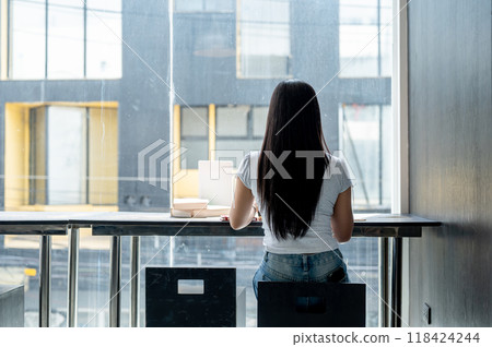 A rear view of an Asian woman sitting at a table by the window, working remotely from a coffee shop. A rear view of an Asian woman sitting at a table by the window, working remotely from a coffee shop. 118424244
