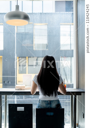 A rear view of an Asian woman sitting at a table by the window, working remotely from a coffee shop. A rear view of an Asian woman sitting at a table by the window, working remotely from a coffee shop. 118424245