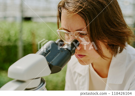 Scientist intently observing sample through microscope in laboratory setting wearing white lab coat and safety glasses, background blurred with laboratory equipment 118426104
