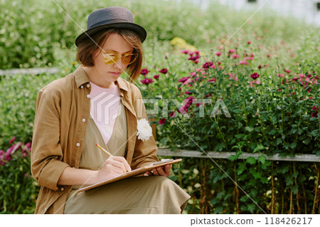 Person sitting in garden and sketching in notebook while surrounded by vibrant flowers. Relaxing in casual clothing and hat, enjoying calm and serene nature environment Person sitting in garden and sketching in notebook while surrounded by vibrant flowers. Relaxing in casual clothing and hat, enjoying calm and serene nature environment 118426217