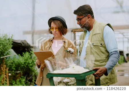 Two people discussing flower arrangements in greenhouse, standing next to lush greenery and flowers, one holding clipboard, another holding flower bouquet Two people discussing flower arrangements in greenhouse, standing next to lush greenery and flowers, one holding clipboard, another holding flower bouquet 118426220