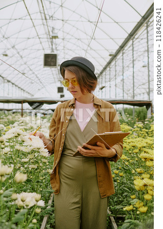 Woman in casual attire analyzing flower growth in greenhouse environment, holding notepad and pen while observing plants around her, yellow flowers dominating the scene 118426241