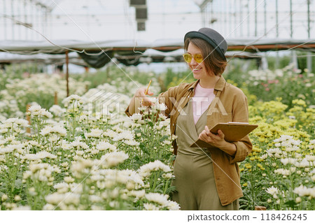 Happy woman wearing hat and sunglasses while inspecting flowers in greenhouse and taking notes. Surrounded by blooming plants and clear sky visible through glass roof Happy woman wearing hat and sunglasses while inspecting flowers in greenhouse and taking notes. Surrounded by blooming plants and clear sky visible through glass roof 118426245