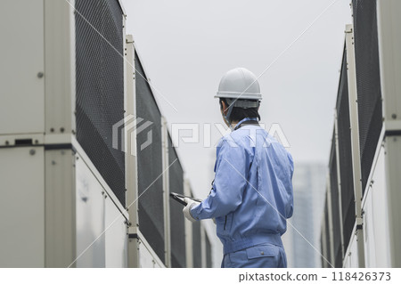 Building maintenance image: A man in work clothes inspecting the outdoor air conditioning unit installed on the roof of a building 118426373