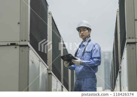 Building maintenance image: A man in work clothes inspecting the outdoor air conditioning unit installed on the roof of a building Building maintenance image: A man in work clothes inspecting the outdoor air conditioning unit installed on the roof of a building 118426374