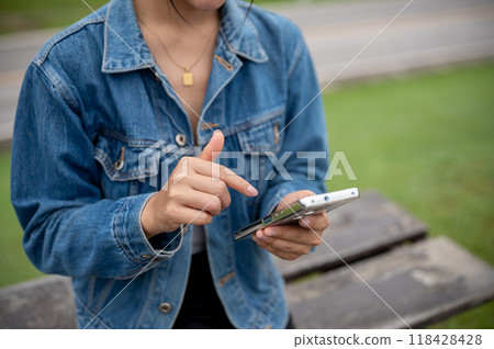 A cropped shot of a woman in a denim jacket using her smartphone while sitting on a bench in a park. 118428428