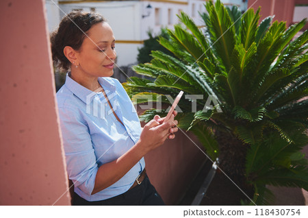 Woman in blue shirt using smartphone outdoors next to a lush green plant on a sunny day 118430754