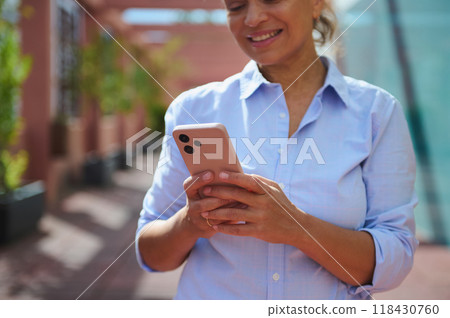 Smiling woman using smartphone outdoors on sunny day, wearing casual blue shirt, surrounded by greenery, enjoying communication and technology Smiling woman using smartphone outdoors on sunny day, wearing casual blue shirt, surrounded by greenery, enjoying communication and technology 118430760