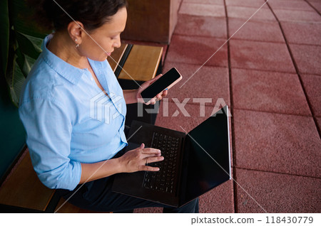 Woman in blue shirt using a laptop and smartphone on a bench outdoors for work and communication 118430779
