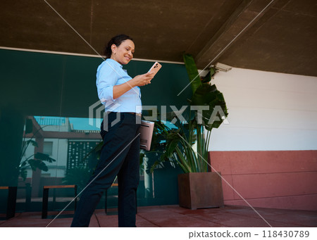 Confident professional woman using smartphone outdoors, smiling and holding laptop in relaxed business setting 118430789