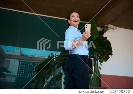 Confident professional woman smiling outdoors while holding a smartphone, showcasing success in business communication and modern technology use Confident professional woman smiling outdoors while holding a smartphone, showcasing success in business communication and modern technology use 118430790