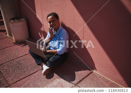 Young woman sitting outdoors using laptop and talking on phone, smiling in sunlight against pink wall Young woman sitting outdoors using laptop and talking on phone, smiling in sunlight against pink wall 118430813