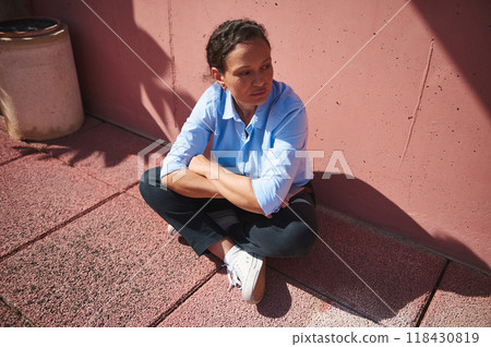 Pensive woman in casual attire sitting against a pink wall in bright sunlight Pensive woman in casual attire sitting against a pink wall in bright sunlight 118430819