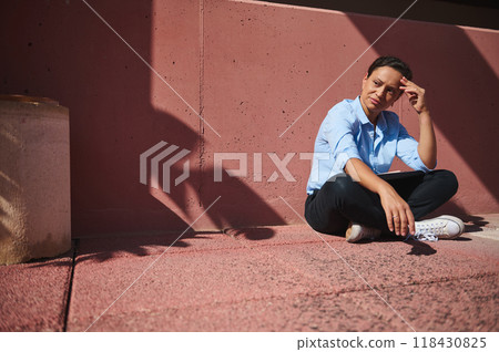 Woman sitting outdoors on a sunny day, thoughtful expression, dressed casually in blue shirt and jeans, relaxing against a concrete wall in an urban setting Woman sitting outdoors on a sunny day, thoughtful expression, dressed casually in blue shirt and jeans, relaxing against a concrete wall in an urban setting 118430825