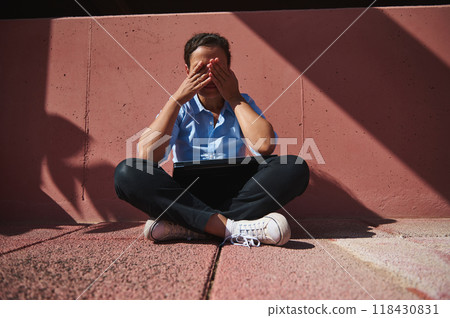 Frustrated person sitting outdoors covering face while using laptop on a sunny day Frustrated person sitting outdoors covering face while using laptop on a sunny day 118430831