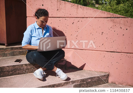 Woman working on a laptop while sitting on outdoor steps with a smartphone beside her, enjoying sunny day and remote work flexibility Woman working on a laptop while sitting on outdoor steps with a smartphone beside her, enjoying sunny day and remote work flexibility 118430837