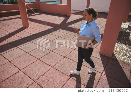 Professional woman walking while using smartphone and holding laptop in modern outdoor setting with red tiles under sunlight 118430932