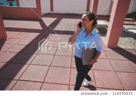 Smiling woman walking outdoors while talking on a smartphone, holding a laptop, conveying a professional and positive mood Smiling woman walking outdoors while talking on a smartphone, holding a laptop, conveying a professional and positive mood 118430935