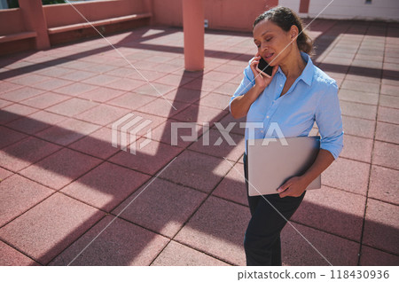 Businesswoman walking outdoors while talking on phone holding laptop under sunlight 118430936