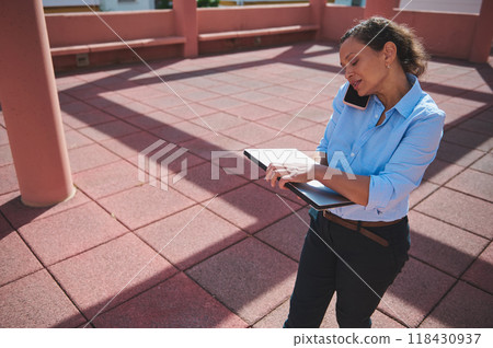 Professional woman multitasking outdoors with a smartphone and clipboard, balancing work and communication efficiently on a sunny day 118430937