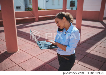 Busy professional woman multitasking with laptop and phone in outdoor workspace environment. 118430938