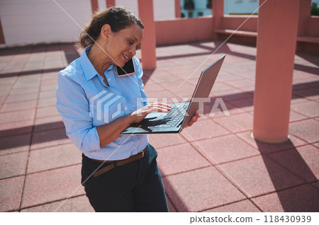 Confident woman multitasking with laptop and phone in outdoor office environment 118430939