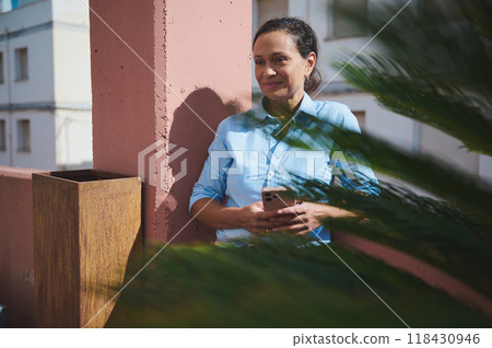 Calm woman in blue shirt holding smartphone outdoors, enjoying peaceful moment and sunshine 118430946