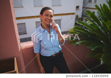 Smiling woman in blue shirt talking on phone outdoors on a sunny day, leaning against a pink wall. 118430949