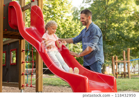 A father stands at the bottom of a red playground slide, smiling and encouraging his young daughter as she slides down. The scene takes place on a sunny summer day 118432950