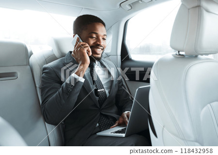 A smiling black businessman in a suit sits in the backseat of a car, working on his laptop while talking on his phone. He appears to be taking a business call and possibly multitasking A smiling black businessman in a suit sits in the backseat of a car, working on his laptop while talking on his phone. He appears to be taking a business call and possibly multitasking 118432958