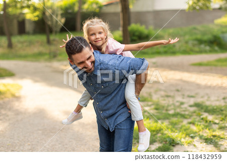 A father gives his daughter a piggyback ride in a park on a sunny day. The father is smiling and the daughter is looking straight ahead, both enjoying the afternoon. A father gives his daughter a piggyback ride in a park on a sunny day. The father is smiling and the daughter is looking straight ahead, both enjoying the afternoon. 118432959