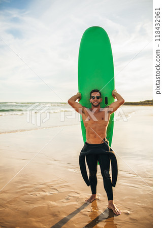 A man in a wetsuit is holding a green surfboard on a sandy beach. He is wearing sunglasses and smiling. The ocean is in the background and the sky is clear. A man in a wetsuit is holding a green surfboard on a sandy beach. He is wearing sunglasses and smiling. The ocean is in the background and the sky is clear. 118432961