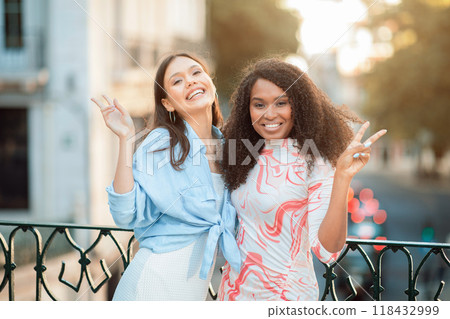 Two women are standing on a city street, smiling and posing with peace signs. They appear to be enjoying their time together as they stand in front of a wrought iron fence. 118432999