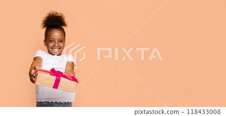 A little African American girl with a big smile holds out a gift wrapped in brown paper and a pink ribbon. She is wearing a white shirt and jeans, and her hair is pulled back in a high ponytail. 118433008