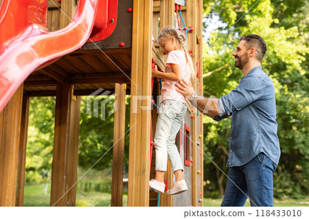 A young girl in pink shirt and jeans is climbing a rope ladder on a wooden playground, while her father encourages her with a smile and a gentle hand on her back. A young girl in pink shirt and jeans is climbing a rope ladder on a wooden playground, while her father encourages her with a smile and a gentle hand on her back. 118433010