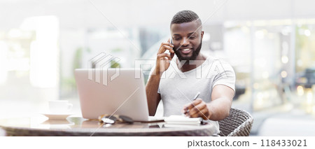 A man with a beard sits at a table outdoors, talking on a phone while working on a laptop and taking notes. 118433021