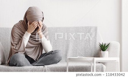 Depression concept. Sad muslim woman in hijab crying on couch at home, covering her face with hands, free space Depression concept. Sad muslim woman in hijab crying on couch at home, covering her face with hands, free space 118433042