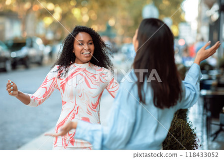 Two multiethnic women meet on a city street on a sunny day. The background is blurred, showing a street with cars and trees. 118433052