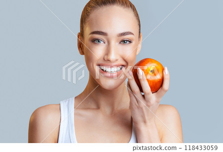A woman with blonde hair and bright blue eyes smiles broadly as she holds a red apple in her hand. The background is a plain grey, allowing her bright smile and the vibrant apple to stand out. 118433059