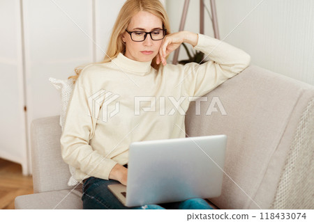 A woman sits on a couch in her living room, wearing glasses and a white sweater, working on a laptop computer. A woman sits on a couch in her living room, wearing glasses and a white sweater, working on a laptop computer. 118433074