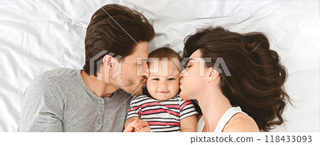A father and mother lay on a white bed and kiss their baby, who is lying between them. The father is on the left side of the image, and the mother is on the right, top view, panorama A father and mother lay on a white bed and kiss their baby, who is lying between them. The father is on the left side of the image, and the mother is on the right, top view, panorama 118433093