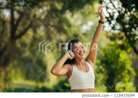 A happy woman with curly hair is dancing in a park while listening to music with her white headphones on. She is wearing a white tank top and her arm is raised in the air, copy space A happy woman with curly hair is dancing in a park while listening to music with her white headphones on. She is wearing a white tank top and her arm is raised in the air, copy space 118433094