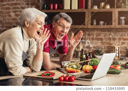 A senior couple, wearing aprons and looking excited, follows a cooking tutorial on a laptop computer in their kitchen. They are surrounded by fresh ingredients A senior couple, wearing aprons and looking excited, follows a cooking tutorial on a laptop computer in their kitchen. They are surrounded by fresh ingredients 118433095