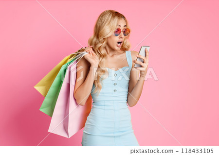 A young woman with long blonde hair is standing in a pink studio, wearing sunglasses and a blue dress. She is holding a smartphone in her right hand, her eyes widened in surprise. A young woman with long blonde hair is standing in a pink studio, wearing sunglasses and a blue dress. She is holding a smartphone in her right hand, her eyes widened in surprise. 118433105